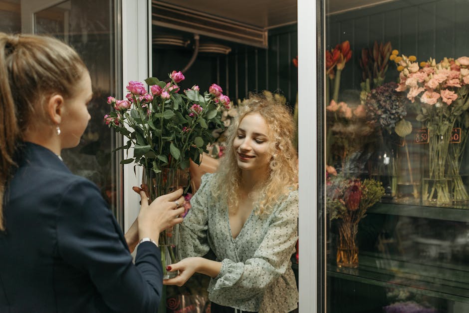 A woman with curly blonde hair, dressed in a light patterned blouse, is receiving a bouquet of pink roses from a florist with brunette hair tied back, wearing a dark blazer, inside a flower shop. The