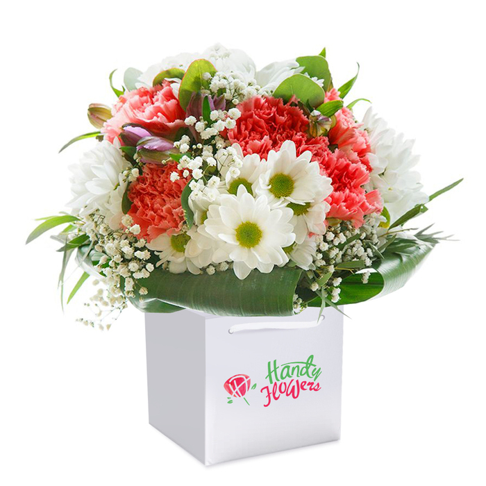 Expertly arranged by Leamouth Florist and standing upright in an elegant white gift bag, this lush bouquet is a showcase of colour, texture, and heartfelt craftsmanship. The composition features generous clusters of coral and red carnations-their distinctive, ruffled petals layering richly over one another-joined by pure white daisies with vibrant chartreuse centers that illuminate the heart of the bouquet. Light sprigs of baby's breath bring an almost weightless softness, weaving through the florals like morning mist rolling over the Thames. On the left, several unopened buds tinged with purple and pink hint at new beginnings and add gentle contrast. Wrapping the arrangement, abundant broad-leaf greenery creates a glossy, deep-green frame, echoing the urban parks and riverbanks nearby. The square white gift bag, practical yet stylish with its sturdy ribbon handles, signals the bouquet's readiness to be gifted-perhaps on a walk through Leamouth's evolving landscapes or delivered to a loved one at home. The pristine white backdrop in the photograph makes each colour and detail pop, accentuating freshness and luxury. This bouquet exudes both joyous exuberance and a quiet sophistication, making it perfect for special celebrations, thoughtful milestones, or simply to bring a touch of Leamouth's floral beauty into any space.