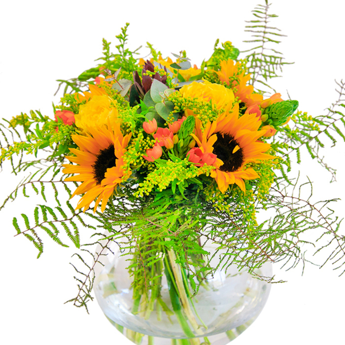 An exuberant celebration of colour and natural texture, this floral centerpiece channels the bright, hopeful feel of a Leamouth morning. Sunflowers, resplendent in their golden attire, take pride of place-each bloom's crisp yellow-orange petals opening outward from a velvety, dark brown core, conjuring memories of sunny walks along the Thames Path. Between these stars, generous yellow roses spiral in densely-layered bouquets, their satiny petals unfolding with luxurious grace. Ample green foliage-delicate ferns, light green filler, and a hint of silvery eucalyptus-spills artfully from the composition, lending it a sense of wild beauty that echoes the lush community gardens nestled around Leamouth. Bright clusters of red Hypericum berries punctuate the bouquet, shining like hidden gems among emerald leaves. All are presented in a perfectly clear, spherical glass vase, its transparency revealing the tangle of healthy stems submerged in sparkling water-a nod to the purity and freshness prized by Leamouth Florist. Soft, natural daylight plays across the scene, casting fluid shadows and enhancing the arrangement's lively disposition. The bouquet's subtle yet inviting fragrance mingles with the air, recalling summer meadows and riverside breezes. This radiant arrangement is a fitting choice for joyful celebrations-birthdays, housewarmings, or simply as a centrepiece to brighten a Leamouth home-bringing a unique sense of place and artistry to every occasion.